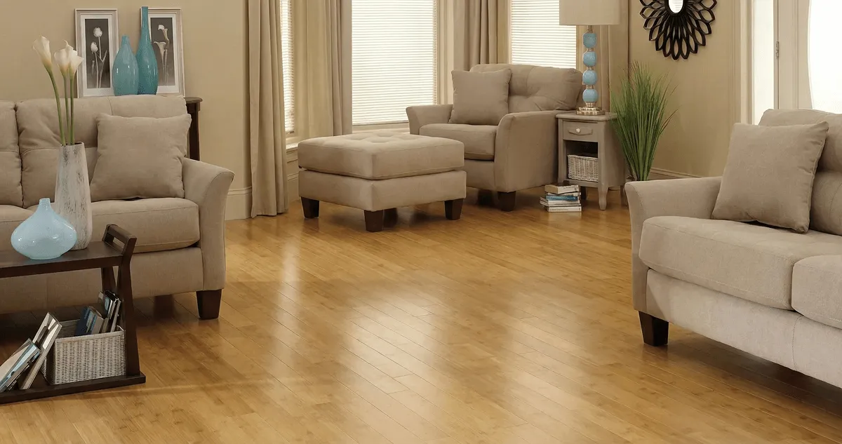 A living room with professionally installed bamboo flooring, beige sofas, a matching ottoman, a side table with a blue lamp, vases, framed pictures, and large windows with blinds and curtains in Irvine, CA